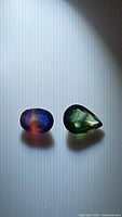 Two natural sapphires photographed on a white surface, showing pear cut green sapphire and oval cut multicolored sapphire with blue and orange hues under natural light.