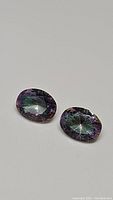 Pair of oval cut Mystic Topaz gemstones with purple rainbow color effect, displayed on a white surface.