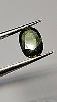 Close-up image of the natural oval cut multicolored sapphire showing a dark green color with yellow sparkles and slight blue streaks held by tweezers.
