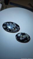 Two oval natural snowflake obsidian cabochons on a white background under light, showing distinctive snowflake patterns and polished surfaces.