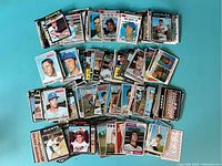 Mixed 1970s baseball trading cards spread out on turquoise background in multiple stacks with visible player photos and teams.