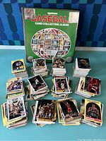 Photo showing green Hygrade Baseball Card Collecting Album binder and multiple stacks of basketball trading cards spread out on a blue surface.
