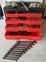 Top view of the black plastic Craftsman toolbox with four red trays extended, showing various wrenches, sockets, and tools inside plus plastic bagged items on top.