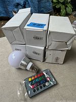 Photo of six white LED bulbs in individual white boxes stacked and one bulb out of box next to a black remote control with colored buttons.
