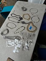 Overview of various silver-tone bracelets, bangle, black bead necklace, and multiple sterling silver earrings packed in clear bags laid out on a table.