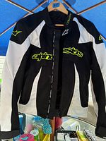 Front view of black, white, and neon yellow Alpinestars jacket hanging on a hanger showing logos on chest and sleeves.