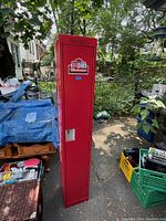 Front view of tall, narrow red metal cabinet with Budweiser NFL logo and handle on door, placed outdoors near clutter.