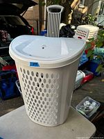 White plastic Sterilite clothes hamper with elliptical holes and a lid placed on a table outdoors with other items in background.