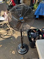 Front view of black Garrison floor fan showing circular grille, fan blades, adjustable stand, base, and attached power cord.
