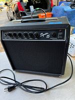 Front view of black Squier 15 guitar amplifier showing control knobs and headphone jack on the top panel, connected power cord visible.