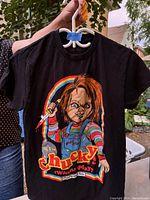 Black T-shirt with colorful Chucky graphic on front, held by person outdoors on white hanger