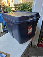 Side view of a large dark gray Rubbermaid Roughneck storage tote with lid placed on a white surface outdoors.