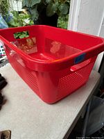 Red plastic laundry basket with perforated sides and integrated handles, placed outdoors with greenery in background.