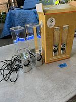 Pair of dancing water speakers with clear water chambers on silver bases alongside original box showing front panel graphic and labeling.
