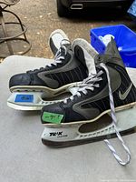 Side view of the two black and grey Nike ice skates with white TUUK custom blade holders sitting on a table, showing laces and profile.