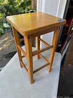 Single wooden stool showing square top and legs with stretchers, natural wood finish, outside photo, some wear visible.