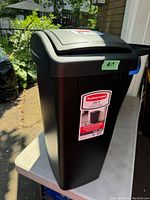 Full view of the Rubbermaid trash can outdoors showing the trash can with a swing top lid, labeled with a price tag.
