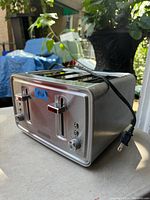 Four slot stainless steel toaster with front lever controls, browning dials, and buttons. Black power cord visible. Light wear on surface.