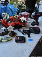 A group photo of power tools including a red and black Black & Decker circular saw, two cordless drills, various battery packs and chargers on a white table outdoors.