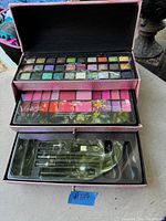 Open makeup case showing three layers of makeup trays with eyeshadows, lip colors, and brush compartments