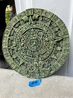 Photo showing a circular vintage Aztec sun disc with intricate carved details and green patina, approximately 10 inches in diameter, held against a white background.