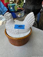 Front and slightly angled view of the vintage hen on nest cookie jar showing the white ceramic hen lid with feather detailing and brown basket-weave base sitting on a table with a blue taped label #85 on the hen.