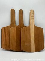 Front view of the three wood cutting/serving boards standing vertically showing their handles and surface finishes, one with 'SUD' carved into handle.