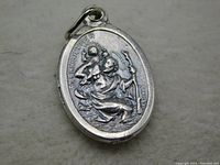 Front side of vintage oval silver tone medal showing Saint Christopher figure carrying a child and holding a staff with the inscription 'St Christopher' and 'Italy' engraved.