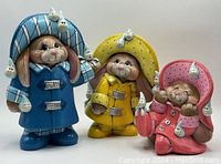 Front view of three ceramic rabbit figurines in colorful raincoats with smiling raindrop decorations.