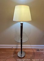 Floor lamp with cream Restoration Hardware fabric shade lit, showing tall slim wood pole, round glass table at midpoint, and heavy metal base.