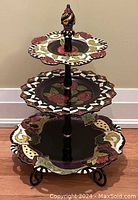 Front view of the large three-tier ceramic serving tray showing scalloped edges, floral patterns, black and white details, and gold accents on plates. The metal stand has a decorative finial and curled feet.