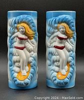 Front view of two ceramic Tikki mugs featuring blonde surfer girls in red bikinis in surfing pose on blue waves.
