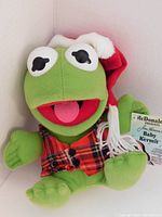 Front view of Baby Kermit plush wearing red and green plaid vest and Santa hat, showing original tags.