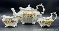 Front view of the set showing the white and gold floral teapot, creamer, and open sugar bowl with scalloped edges and ornate handle details.