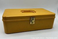 Closed dark yellow rectangular plastic box with metal latch and recessed handle on lid.