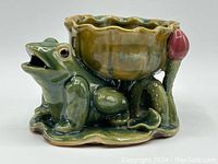 Side view of shiny green glazed ceramic frog figurine on lily pad with brownish scalloped bowl and pink flower decoration.