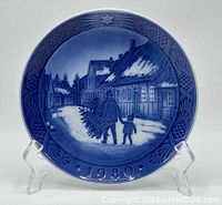 Front view of the blue and white porcelain plate showing a snowy village street with two figures carrying a Christmas tree as evening sets in. The border is embossed with stars and pine cones and the year 1980 is visible at the bottom.