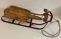 Doll sleigh from angled side view showing wooden slat top, red metal frame, and rope handle.