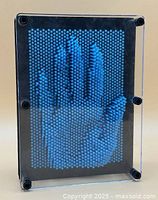 Front view showing blue pins pushed to form a hand impression within a clear plastic frame with four corner screws and black backing.