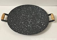Top-down view of round grill pan showing black speckled surface and wooden handles on each side.