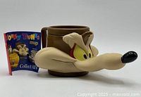 Front angle showing the 3D face design of Wile E. Coyote on the brown plastic mug, with paper insert beside it.