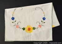 White cotton table runner laid flat showing full embroidery design with yellow, pink, blue flowers and green stitched edge.