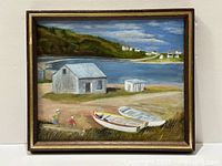 Framed coastal landscape painting showing boats on shore, buildings, hills and water.