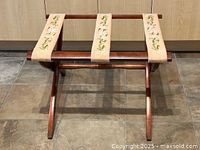 Front view of the wooden luggage rack showing three floral needlepoint straps and wood construction.