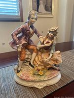 Front angled view of ceramic figurine showing man seated in a purple coat, woman in orange dress, floral decorated base.