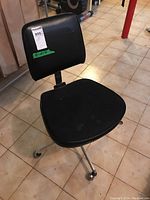 Front and top view of the black office chair showing vinyl backrest and cloth seat upholstery with metal base on caster wheels on tile floor.