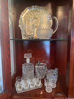 Full view of glass pitcher with handle, decanter with stopper, two ice buckets with tongs, six shot glasses on rectangular tray, all on wooden shelf.
