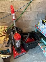 Red Dirt Devil Featherlite upright vacuum with coiled power cord and attached handle, standing upright on floor next to other household items.
