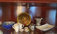 Wide view of all items including espresso cups, stovetop coffee maker, creamer, ceramic bowls, decorative plate, sushi dishes, and serving tray inside wooden cabinet.