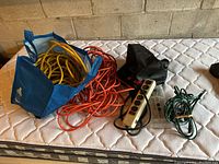 Photo shows two heavy duty extension cords, one orange loosely coiled and one yellow in a blue reusable bag, along with two power bars and a black pouch on a mattress